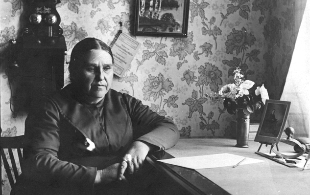 Mustavalkoinen valokuva, jossa nainen istuu pöydän ääressä vanhanaikaisessa asussa.