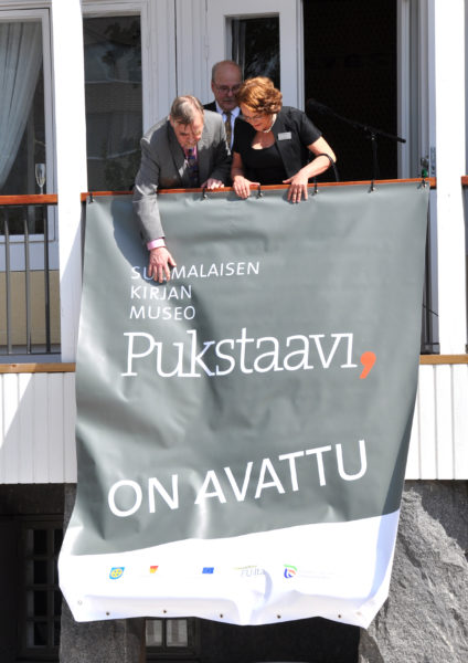 Kolme henkilöä seisoo Pukstaavin terassilla ja avaa banderollia. Banderollissa lukee Suomalaisen kirjan museo Pukstaavi on avattu.